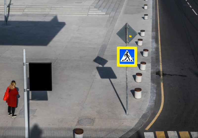 A woman in a red coat walking along a paved street, surrounded by bollards, road signs, strong shadows, and geometric patterns viewed from above.