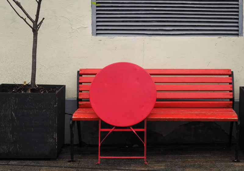 A bright red circular table positioned in front of a red bench, with a small tree in a black planter and a wall vent completing the minimalist urban scene.