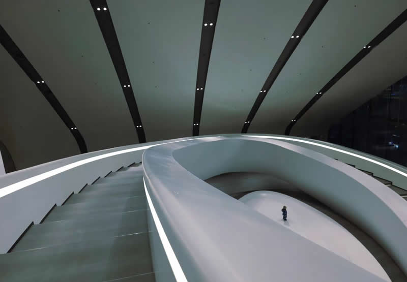 A tiny person standing inside a futuristic white architectural space with sweeping curved ramps, glowing edges, and dark linear ceiling beams.