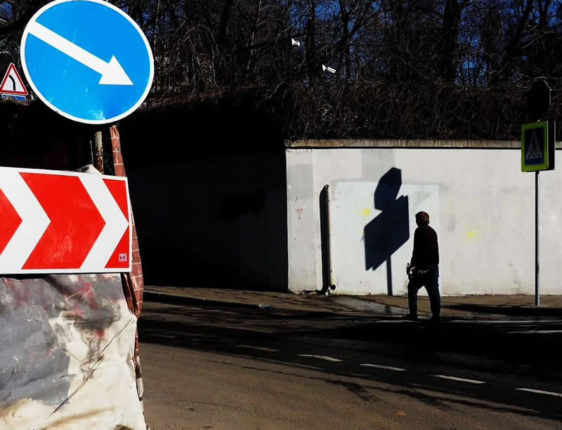 A person walking along a street beneath a large white wall, framed by bold blue and red road signs and strong geometric shadows.