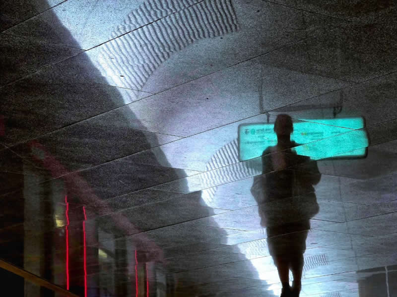 A silhouetted person walking beneath a reflective ceiling, surrounded by geometric light patterns, reflections, and a glowing turquoise sign in a modern urban interior.