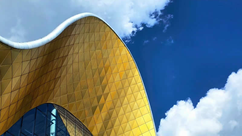 A sweeping golden architectural roof with geometric panels rising against a vivid blue sky with white clouds.