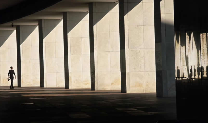 A solitary person walking beneath a large modern structure, framed by tall stone columns, strong geometric shadows, and repeating architectural forms.