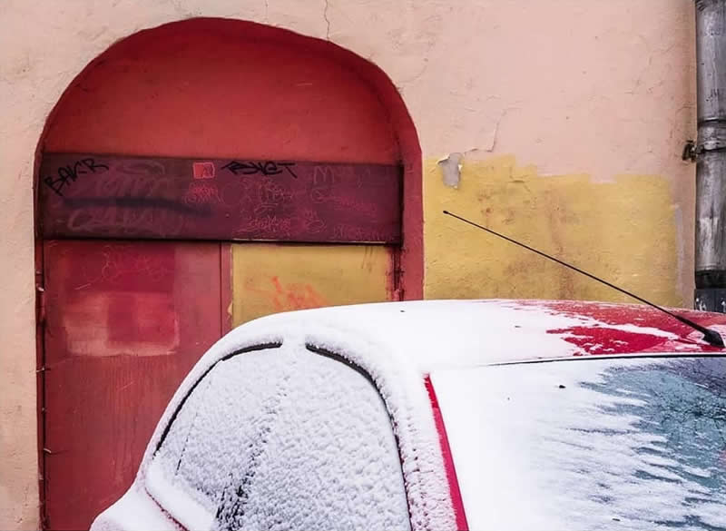 A snow-covered white car parked in front of a weathered red and yellow doorway, creating a colorful urban winter composition.
