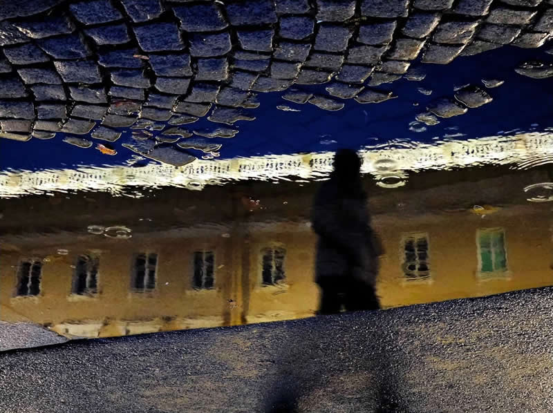 A dark silhouette reflected in a puddle, with a yellow building, windows, blue sky, and cobblestone pavement creating a surreal inverted urban scene.