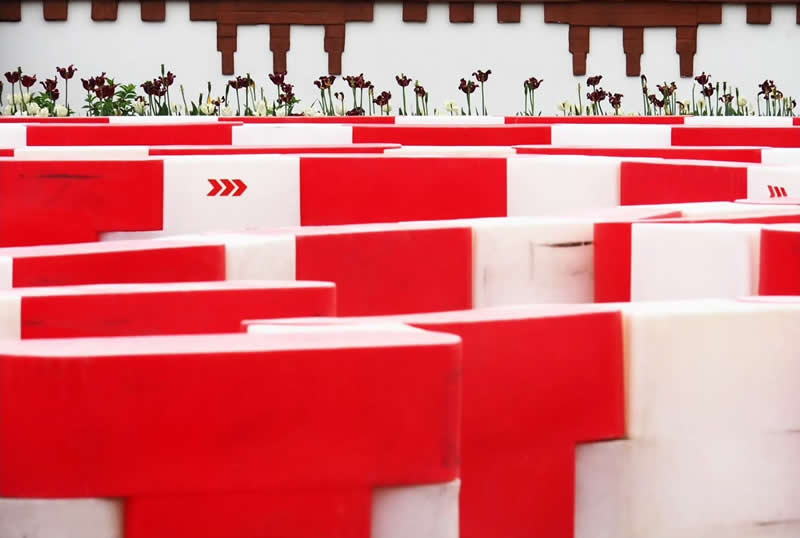 A maze-like arrangement of red and white geometric barriers, with small dark flowers lining the background beneath a white wall.
