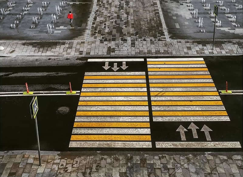 High-angle view of a city street featuring yellow-and-white crosswalks, directional arrows, patterned pavement, traffic signs, and a small person wearing red.