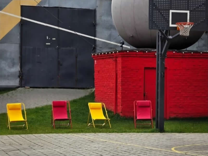 Four colorful folding chairs on grass beneath a basketball hoop, with a vivid red building, gray walls, and geometric court markings in the foreground.