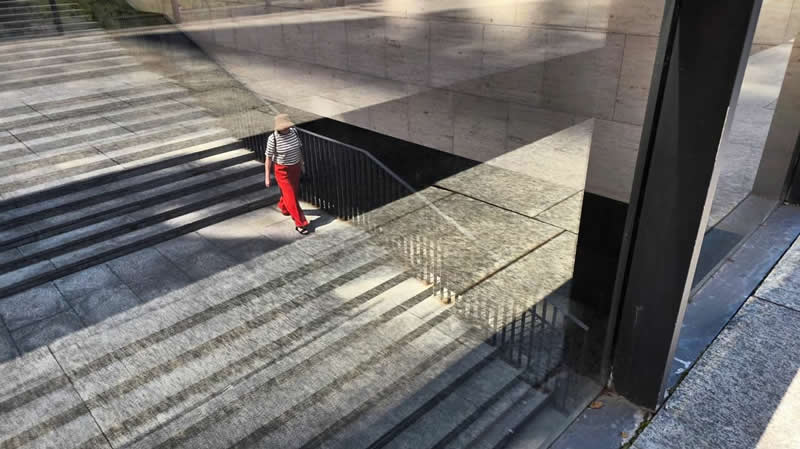 A person wearing red pants walks through a modern concrete space with stairways, railings, strong shadows, and reflective surfaces creating layered geometric patterns.