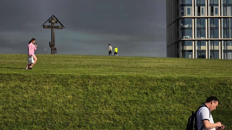 People scattered across a broad grassy slope beneath dark clouds, with a modern glass building on the right and a wooden signpost near the horizon.