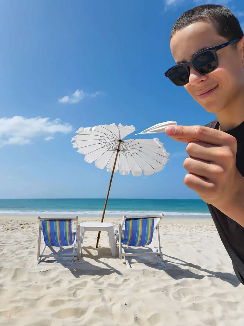 A young man wearing sunglasses appears to pinch a small white beach umbrella between his fingers through forced perspective, with colorful beach chairs, white sand, and blue ocean in the background.