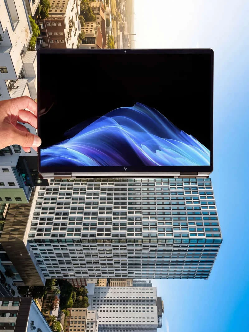 A hand holds a laptop in front of tall city buildings, creating a forced-perspective illusion that makes the computer appear enormous against the urban skyline.