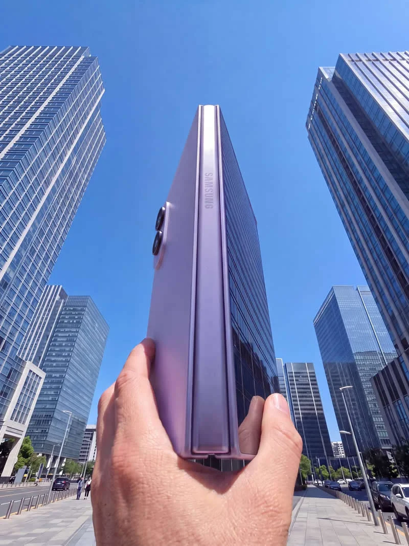 A hand holds a purple Samsung smartphone vertically between tall glass skyscrapers against a clear blue sky, creating a forced-perspective illusion that makes the phone appear as tall as the buildings.