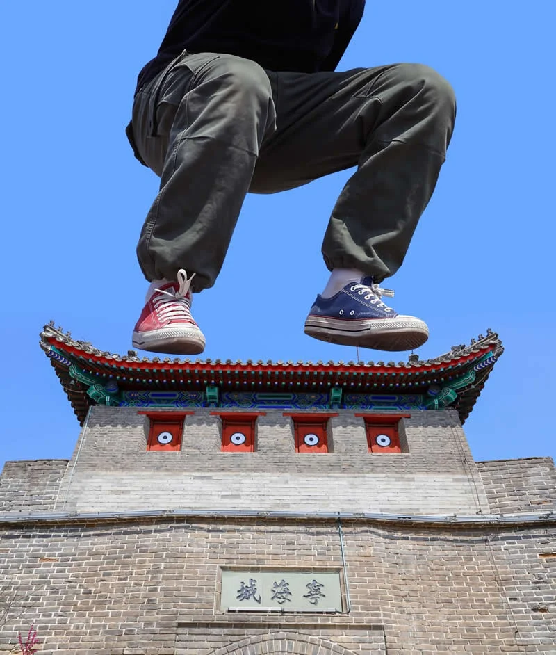 A person wearing mismatched red and blue sneakers appears to squat above the ornate roof of a traditional Chinese-style stone gate against a clear blue sky, creating a forced-perspective illusion.