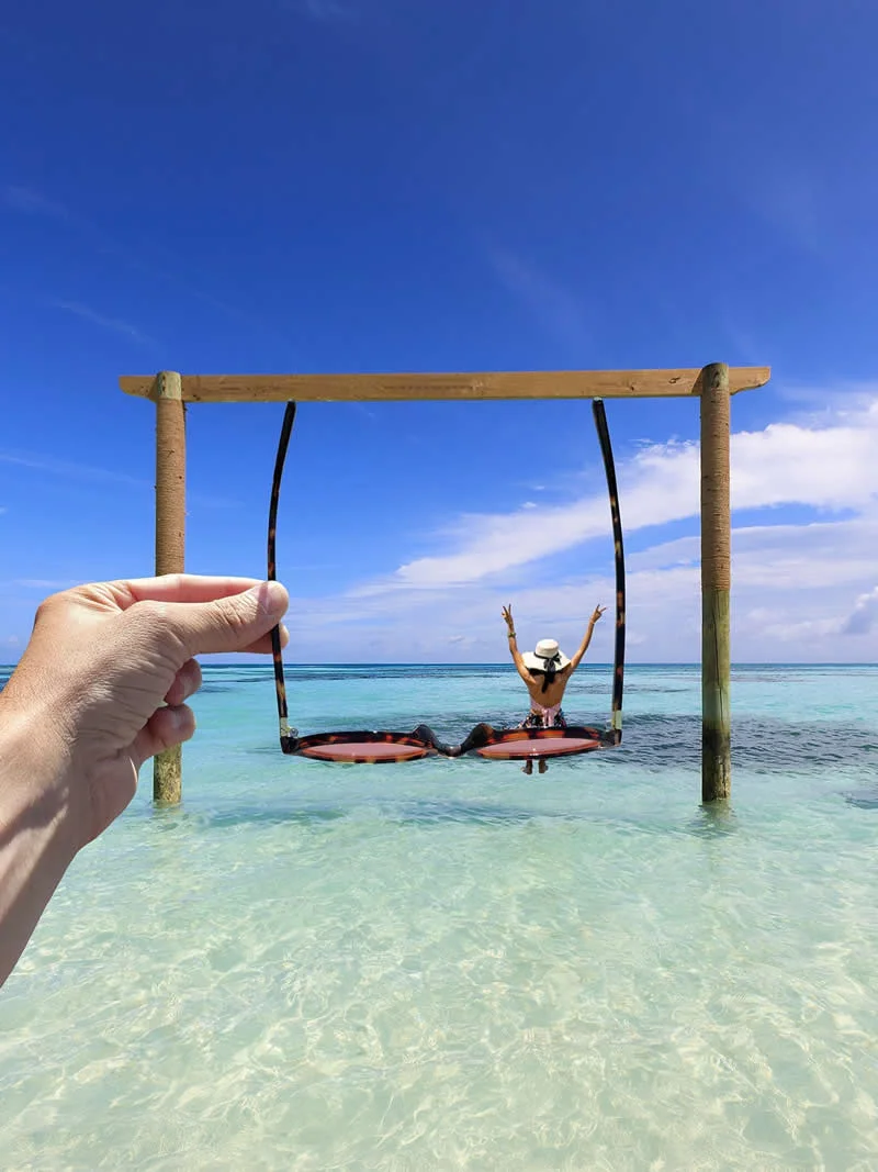 A hand holds oversized sunglasses in front of a woman posing with raised arms in clear turquoise seawater, framed by the glasses beneath a wooden beach structure and a bright blue sky.
