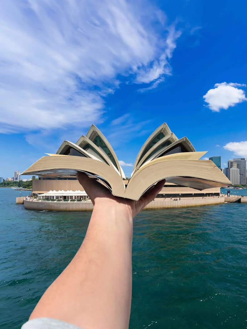 A hand holds an open book in front of the Sydney Opera House, aligning its pages with the building&rsquo;s sail-shaped roofs to create a forced-perspective illusion above Sydney Harbour.