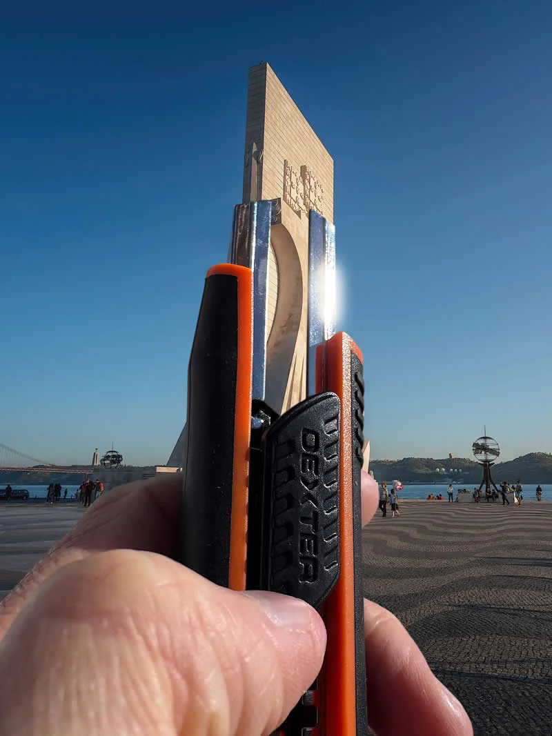 A hand holds an orange-and-black utility knife in front of a tall waterfront monument, aligning the tool with its structure to create a forced-perspective illusion against a clear blue sky.