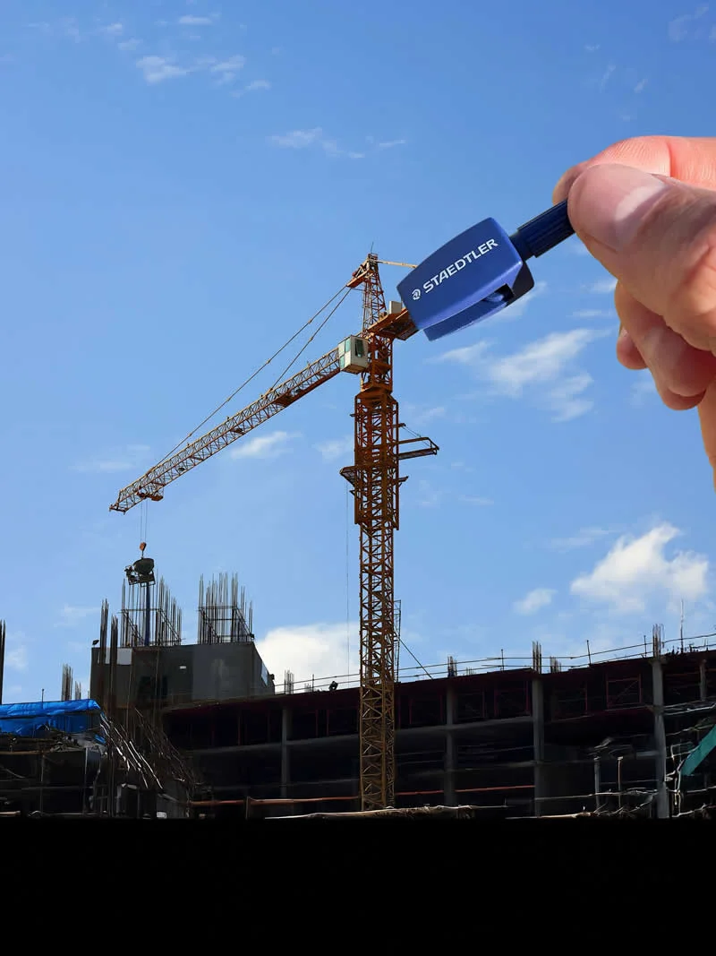A hand holds a blue STAEDTLER pencil sharpener beside a tall construction crane, creating a forced-perspective illusion that makes the sharpener appear to grip the crane&rsquo;s upper structure against a bright blue sky.