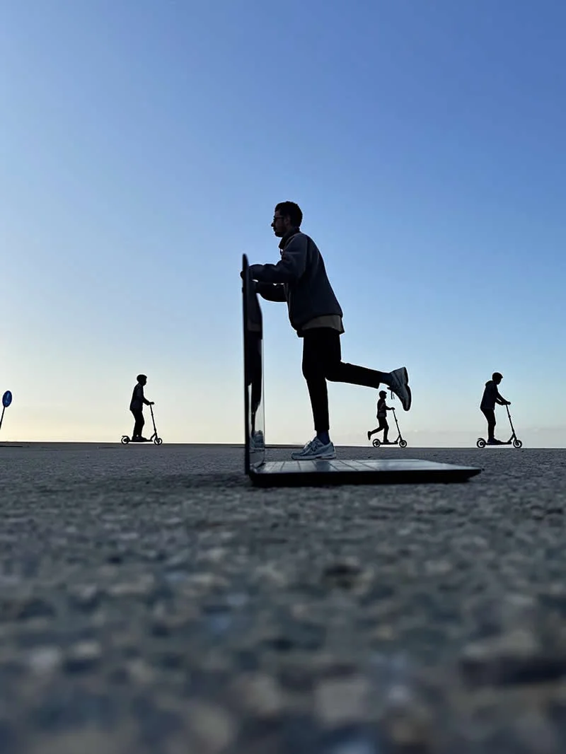A man appears to balance on the screen of an oversized laptop while three people ride electric scooters in the background, creating a forced-perspective illusion against a clear blue sky.