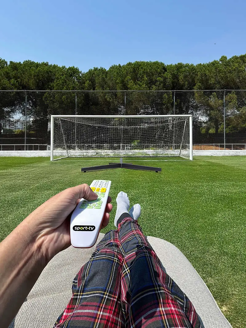 A person wearing plaid trousers and white socks reclines on a chair beside a football field, holding a television remote aimed at an empty soccer goal, creating the illusion of controlling the game from a comfortable seat.