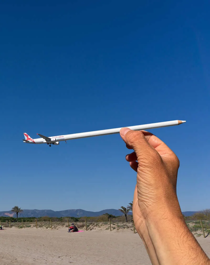 A hand holds a white pencil beneath an Air Alg&eacute;rie airplane flying across a clear blue sky, creating the illusion that the pencil supports the aircraft above a sandy beach with distant mountains and palm trees.