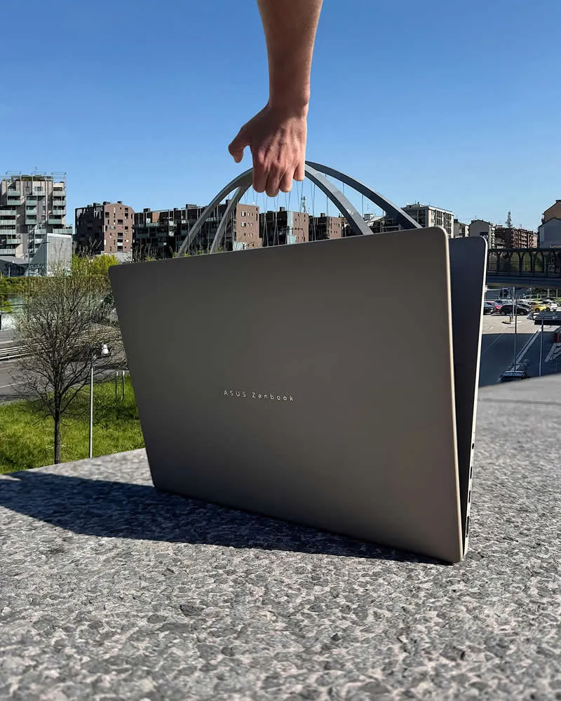A hand reaches down toward an ASUS Zenbook laptop positioned on a stone ledge, aligned with the arch of a modern bridge behind it to create the illusion of lifting the laptop using the bridge, with city buildings and a clear blue sky in the background.