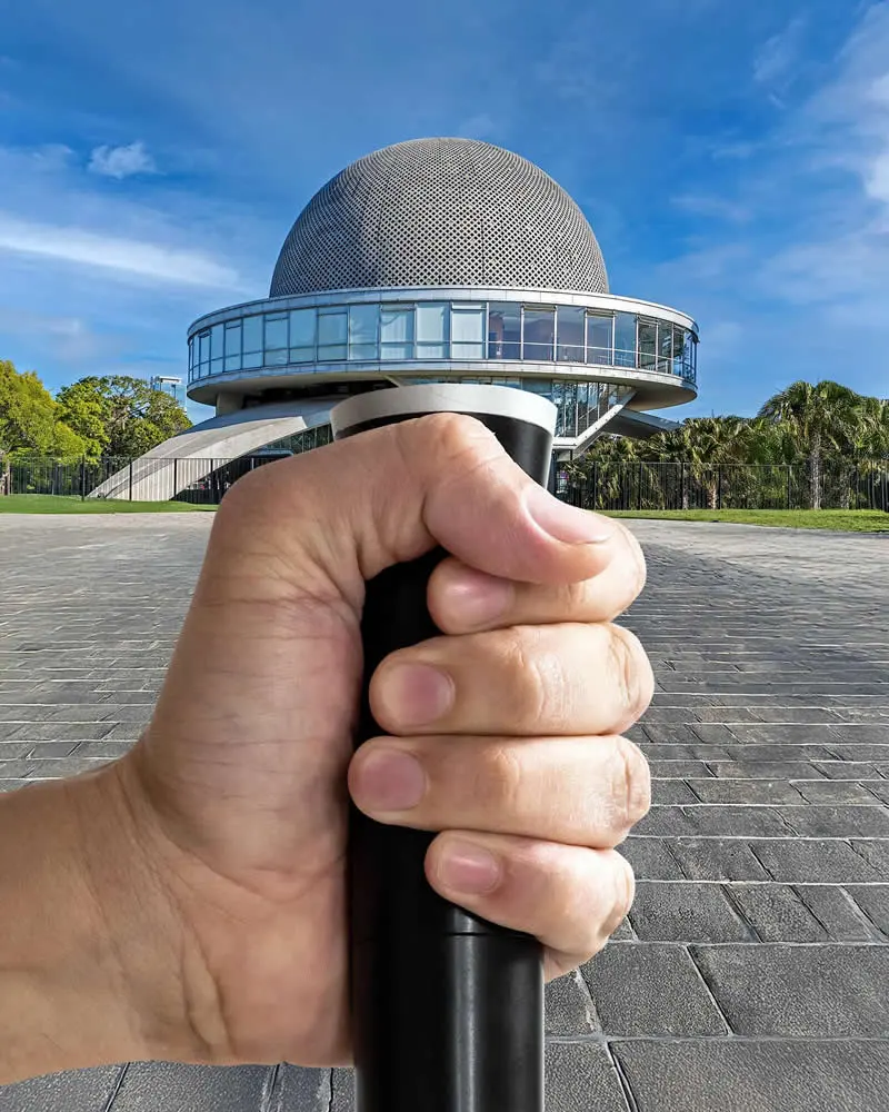 A close-up of a hand gripping a black handle, positioned to make it appear as though it supports a futuristic circular building topped with a perforated dome. The modern glass structure stands against a bright blue sky with wispy clouds and green trees.