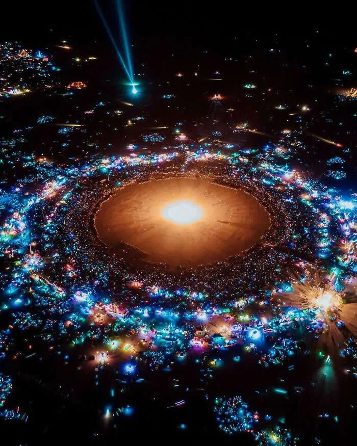 Aerial night view of Burning Man with a glowing central light surrounded by colorful illuminated camps, resembling a solar system in the dark desert sky.