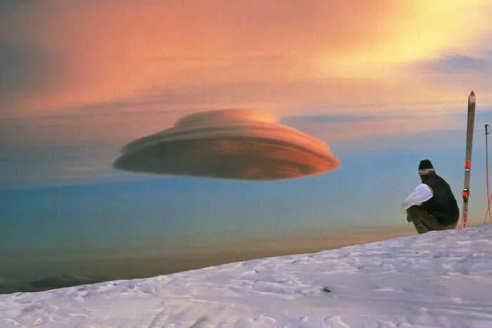 Saucer-shaped lenticular cloud resembling a UFO floats above a snowy mountain beneath a colorful sunset sky, with a person crouching beside a tall piece of equipment.