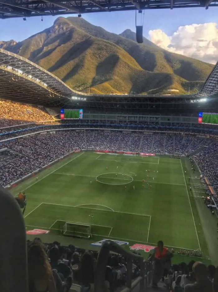 Football stadium packed with spectators, featuring a green pitch and towering mountain peaks rising dramatically above the open roof under a blue sky.