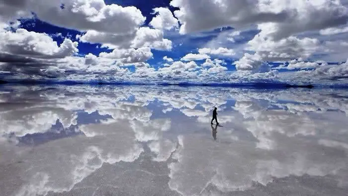 A lone person walks across Bolivia's Salar de Uyuni salt flat, where dramatic white clouds and a deep blue sky create a stunning mirror reflection on the wet surface.