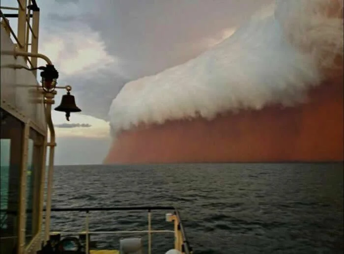 Massive reddish dust storm with a towering white cloud rolls across the sea off Australia, viewed from a boat with its railing and bell visible in the foreground.