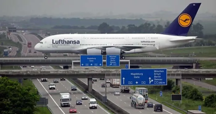 Lufthansa Airbus A380 appears to cross a German autobahn above cars and trucks, with highway signs and green countryside surrounding Leipzig Airport.