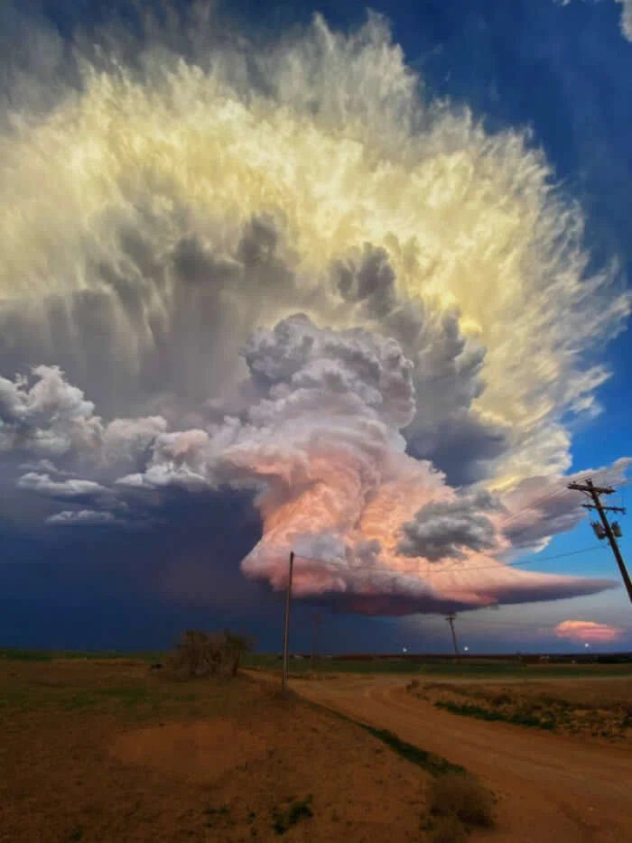 Massive towering storm cloud with swirling gray, pink, and golden formations spreads across a blue sky above a rural dirt road, open fields, and utility poles.