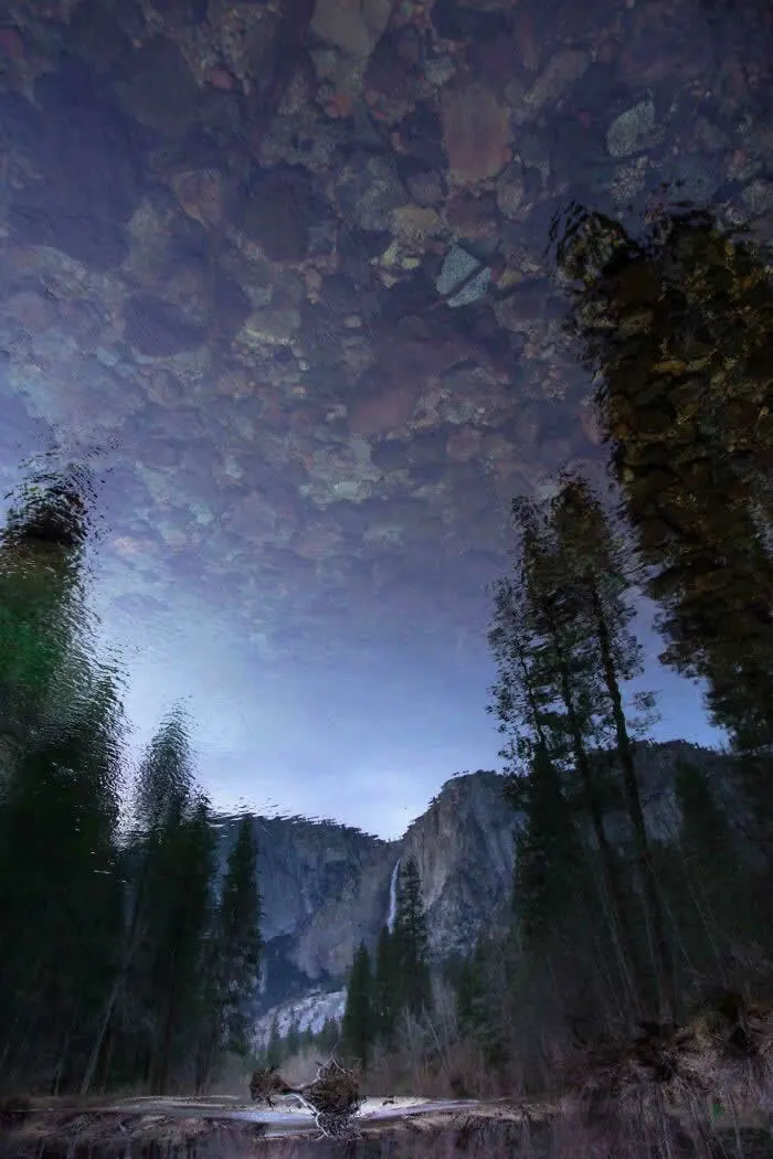 Rocky river reflects towering cliffs, tall pine trees, and the sky in Yosemite Valley, creating a surreal landscape with a mirror-like surface and subtle ripples.