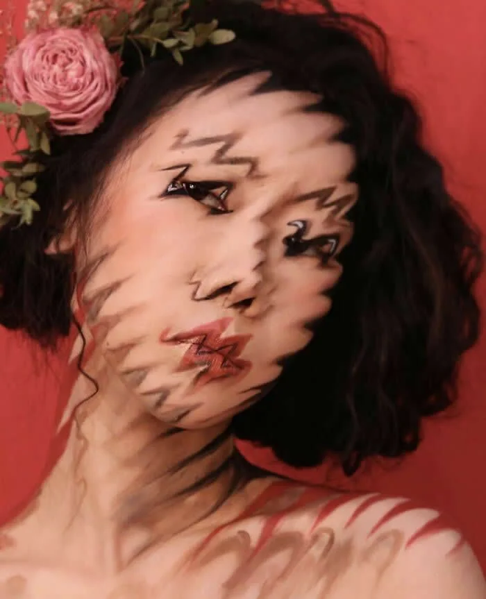 Woman with short dark hair and a pink flower wears intricate zigzag makeup across her face, creating a fragmented optical illusion against a coral-red background.