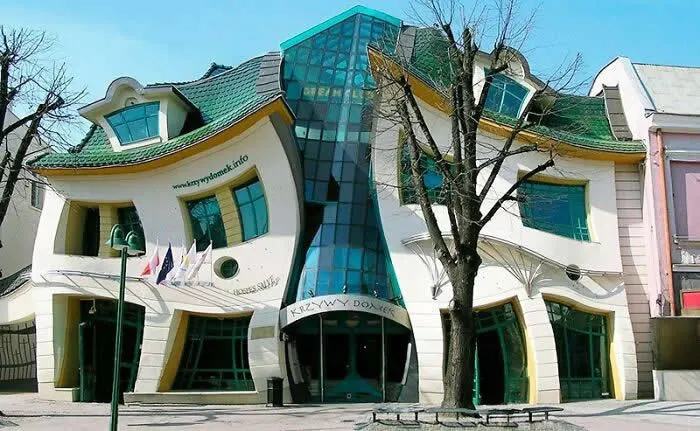 Krzywy Domek, Poland&rsquo;s famous Crooked House, features warped white walls, curved green-framed windows, a wavy roof, and a striking blue glass entrance.