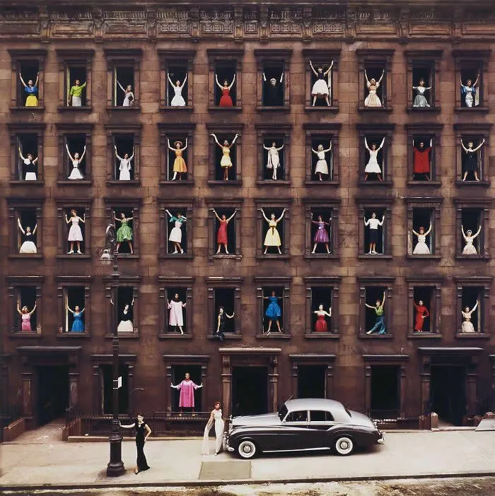 Vintage black-and-white-era street scene showing dozens of people in colorful outfits posing with raised arms from rows of windows in a historic building, with a classic car parked outside.
