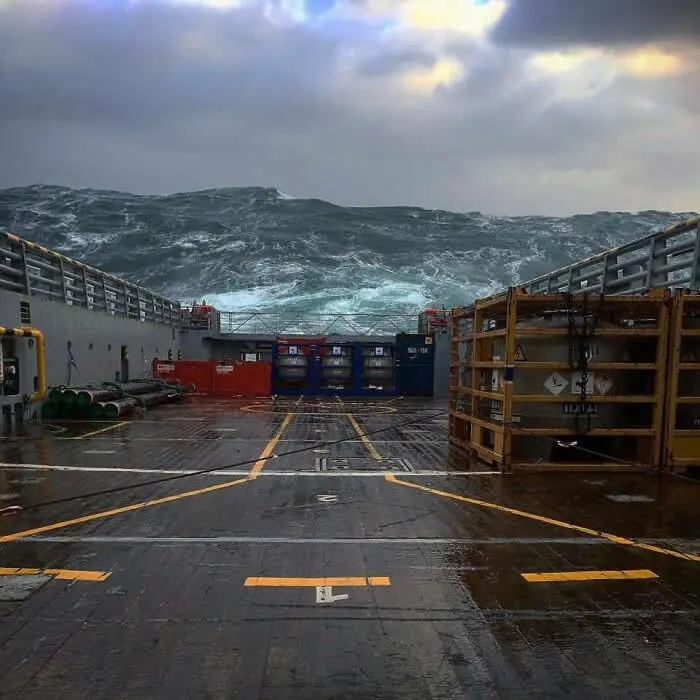Massive dark waves rise behind the wet industrial deck of a ship in the North Sea, with equipment containers, metal railings, and storm clouds in the background.