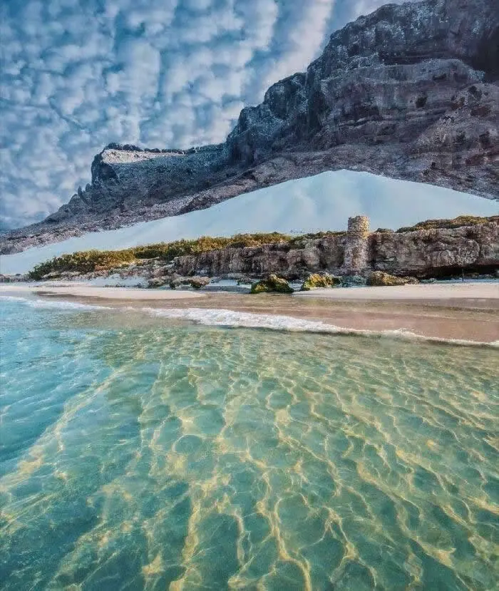 Rugged dark mountains with unusually pale blue sections rise above a rocky shoreline and clear turquoise water, beneath a sky filled with textured white clouds.