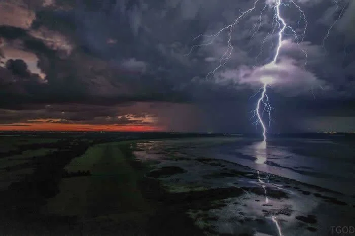 Bright lightning bolt strikes the water beneath dark storm clouds, captured from a drone at sunset, with an orange glow along the horizon and reflections on the wet landscape.