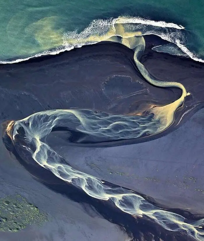 Aerial view of pale winding streams flowing across dark volcanic sand toward turquoise ocean water, creating elegant, abstract patterns along a rugged coastline.