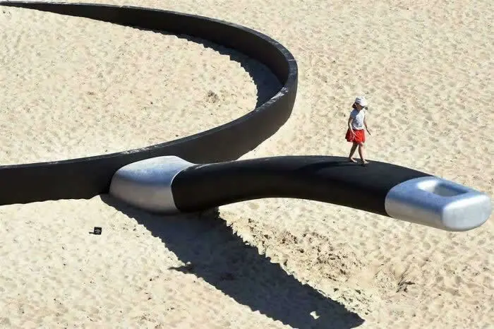 Large black spoon-shaped sculpture lies across a sandy beach in Sydney, Australia, with a woman in a red skirt walking along its curved handle.