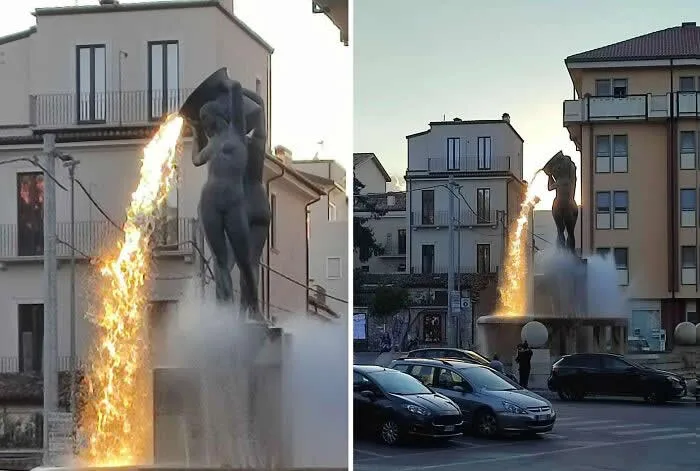 Outdoor fountain featuring a dark statue that appears to pour a bright stream of fire into the water below, surrounded by residential buildings and parked cars.
