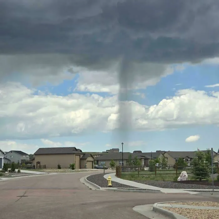 A narrow column of rain falls from a dark storm cloud onto a single area of a suburban neighborhood, while blue skies and white clouds remain visible nearby.