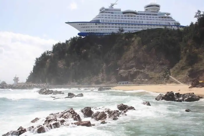 Giant white cruise ship-shaped hotel perched on a forested coastal cliff in South Korea, overlooking a rocky beach and waves crashing against the shore.
