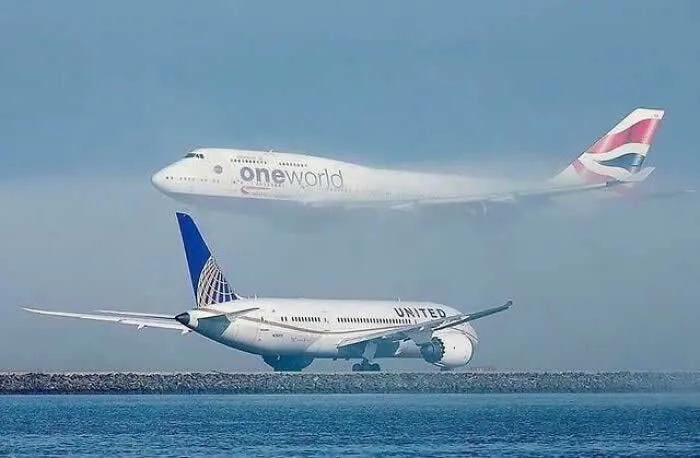 British Airways Boeing 747 flying above a United Airlines passenger jet near the runway, with blue ocean water in the foreground and a clear sky behind them.