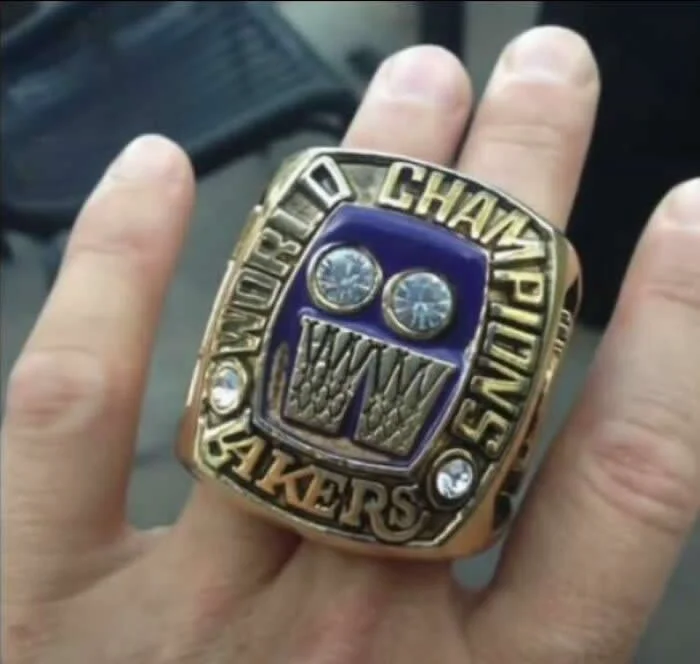 Oversized gold Los Angeles Lakers championship ring with a purple center, decorative lettering, and sparkling stones worn on an ordinary person&rsquo;s hand.