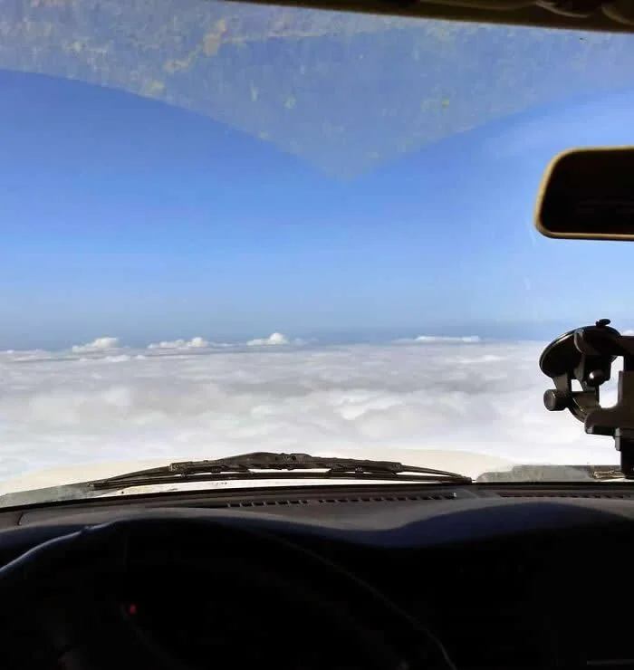 View from inside a parked car at a mountain summit, looking through the windshield at a vast sea of white clouds beneath a clear blue sky, creating the illusion of flying above the clouds.