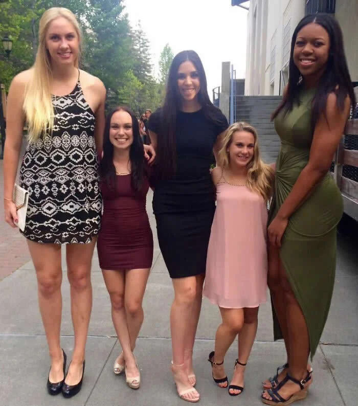 Five women of noticeably different heights pose together on a sidewalk outside a building, wearing dresses in black, burgundy, pink, and green.